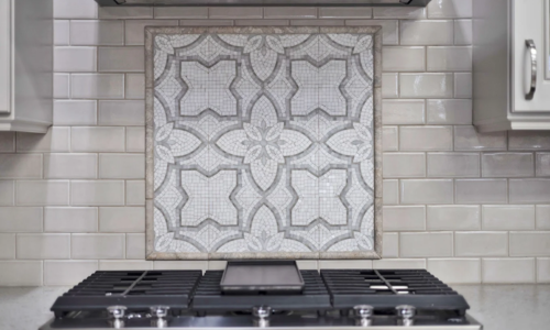 Decorative gray patterned backsplash tile panel with geometric and leaf motifs mounted on a light brick backsplash above a black gas cooktop with metal burners and knobs.